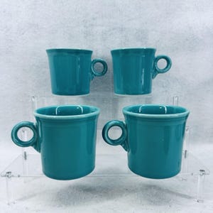 Könnte beinhalten: Ein Set aus vier passenden, blaugrünen Keramikbechern mit glatter, glänzender Oberfläche. Jeder Becher verfügt über einen zylindrischen Körper, einen breiten, kreisförmigen Henkel und ein dezentes Ringdetail am oberen Rand.