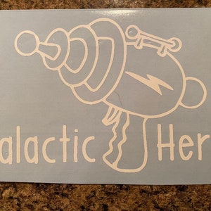 May include: A white vinyl decal featuring a cartoon ray gun with a lightning bolt and the words "Galactic Hero". The design is on a light blue background.