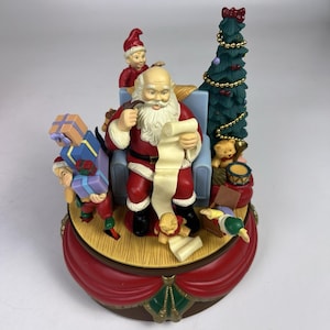 May include: A Christmas-themed figurine featuring Santa Claus seated in a blue armchair, reading a list. Elves are carrying gifts. A Christmas tree and other toys are also present. The base is red with green accents.