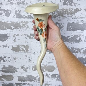 May include: A cream-colored ceramic vase with a flared top and a curved, horn-like stem. The vase is adorned with hand-painted orange flowers and green leaves. The vase is approximately 12 inches tall.