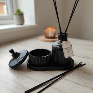 Op de afbeelding: Een zwarte keramische diffuser set met een geribbeld ontwerp. De set bevat een fles met rietjes, een klein schaaltje, een deksel en een dienblad. Een label op de fles luidt "Handmade Ceramic Diffuser Set."