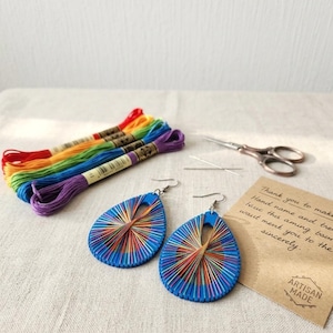 以下が含まれることがあります： 複雑なマルチカラーの糸刺繍が施された青いティアドロップ型のイヤリング1組。虹色の刺繍糸5束、小さな金属製のハサミ、縫い針2本、テキストが記載されたベージュのカードが添えられています。