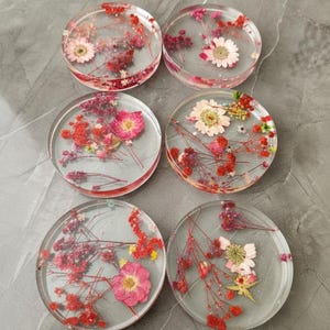 May include: Six clear resin coasters with dried flowers embedded inside. The coasters feature a variety of pink, red, and white flowers and greenery. The coasters are round and appear to be approximately 4 inches in diameter.