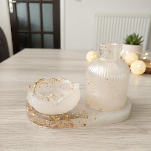 Könnte beinhalten: Ein dekoratives Harz-Set mit einer kleinen, klaren, gerippten Flasche und einem blütenförmigen Kerzenhalter. Das Set ist transparent mit goldenen Glitzerakzenten und steht auf einer passenden ovalen Basis.