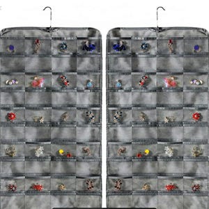 Puede incluir: Dos organizadores de joyas grises colgantes con múltiples bolsillos transparentes, cada uno con varios pendientes, anillos y broches. Las joyas son de varios colores, incluyendo azul, rojo y dorado. Los organizadores cuelgan de un gancho de metal.