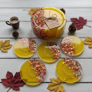 Op de afbeelding: Een handgemaakte Autumn Coaster Set met zes gele hars onderzetters, elk met ingebedde rode en gouden glitter in een bladerenpatroon. De set is vastgebonden met touw en bevat een kleine koffiekop en decoratieve bladeren.