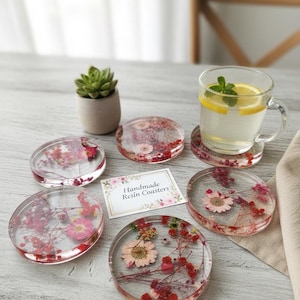 May include: Clear resin coasters with dried flowers in shades of pink and red. A glass of water with lemon and mint sits on one coaster. A small succulent plant in a gray pot is also present. The coasters are handmade and the text on the card reads "Handmade Resin Coasters".