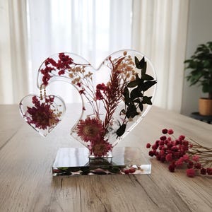 May include: A clear resin heart-shaped sculpture with dried flowers in shades of red, white, and black. The sculpture has a smaller heart attached and sits on a rectangular base. A bouquet of red flowers is on the table.