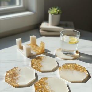 May include: A set of white and gold resin coasters with a matching holder. The coasters are octagonal, with gold leaf accents on a white background. A glass of water with a lemon slice sits on one coaster.