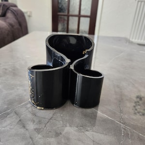 May include: A black desk tidy with three compartments and a glossy finish. The organiser has a distinctive, curved design and gold leaf detailing. It is placed on a grey, marble-effect surface.