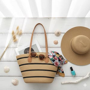 Op de afbeelding: Een beige strooien draagtas met zwarte horizontale strepen, bruine leren handvatten en een zonnebril. Een breedgerande strohoed, een bloemen sjaal en turquoise nagellak suggereren een strand- of zomerthema. De tas bevat een tijdschrift.
