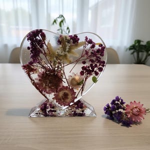 Pode incluir: Uma escultura em forma de coração de resina transparente com flores secas em tons de roxo, rosa e branco. A escultura assenta numa base facetada. Um pequeno arranjo de flores roxas e rosas está ao lado. Decoração de casa.