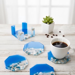 May include: Set of five ocean-themed resin coasters. Each coaster features a blue and white design with embedded pebbles and shells. A white coffee cup sits on one coaster. A blue coaster holder is also present.