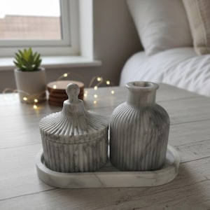 Op de afbeelding: Een decoratieve set met marmerlook, bestaande uit een pot met deksel en een kleine vaas, beide met verticale geribbelde details, op een ovaal dienblad. De set is in grijstinten en wit, wat een minimalistische esthetiek creëert. De tray is ongeveer 25 cm lang.