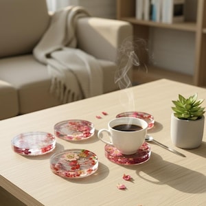 May include: A set of five clear resin coasters with embedded red and yellow flowers. One coaster holds a white cup of coffee, with steam rising. A small succulent plant in a gray pot sits nearby on a light wood table.