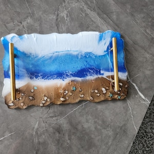 Puede incluir: Bandeja rectangular de resina con diseño de escena de playa. La bandeja presenta un océano blanco y azul, una orilla arenosa con conchas marinas y estrellas de mar, y asas doradas. Mide aproximadamente 30 cm de largo y 15 cm de ancho.