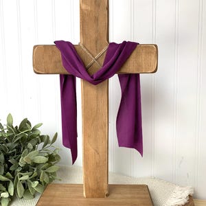 May include: A wooden cross with a purple cloth draped over it. The cross is made of light brown wood and stands on a rectangular base. The purple cloth hangs down on either side of the cross.
