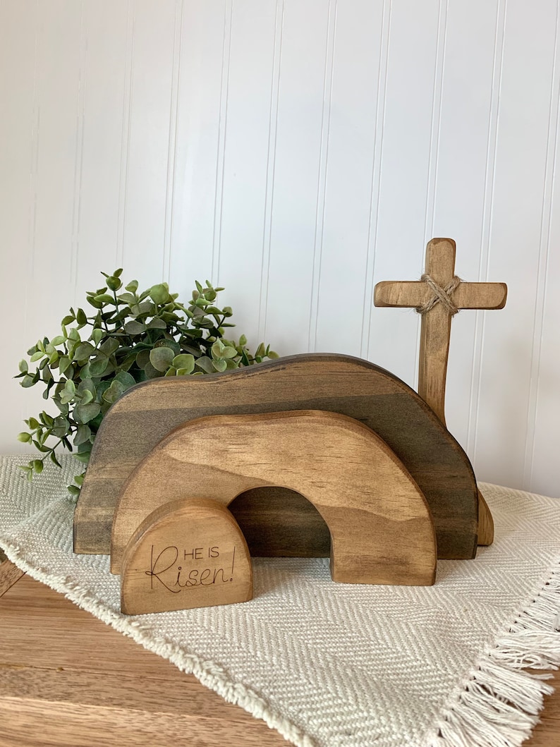 May include: Wooden Easter cross and tomb with the text "He is Risen!" engraved on the front. The tomb is made of three stacked arches, and the cross is tied with twine.