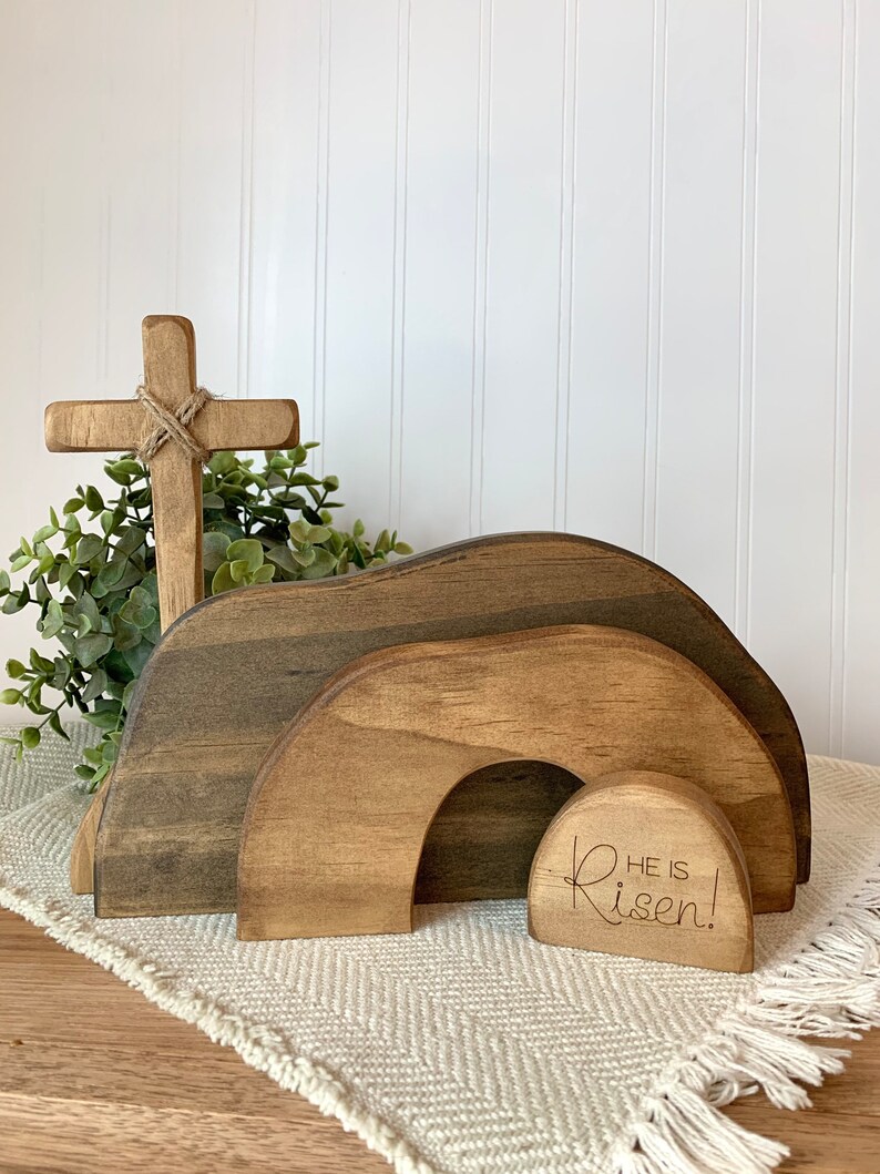 May include: Wooden Easter tomb with a cross and a small sign that reads "He is Risen!" The tomb is made of three pieces of wood, each with a different wood grain. The cross is made of a single piece of wood and has a rope tied around it. The sign is made of a single piece of wood and has the words "He is Risen!" engraved on it.