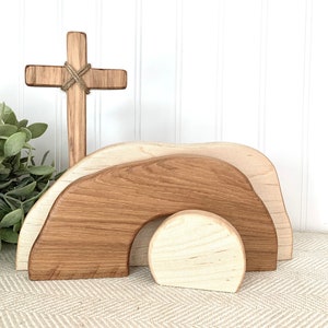 May include: Wooden Easter tomb set with a cross. The tomb is made of three pieces, a large brown piece, a smaller brown piece, and a round white piece. The cross is made of brown wood and has a piece of twine tied around it.
