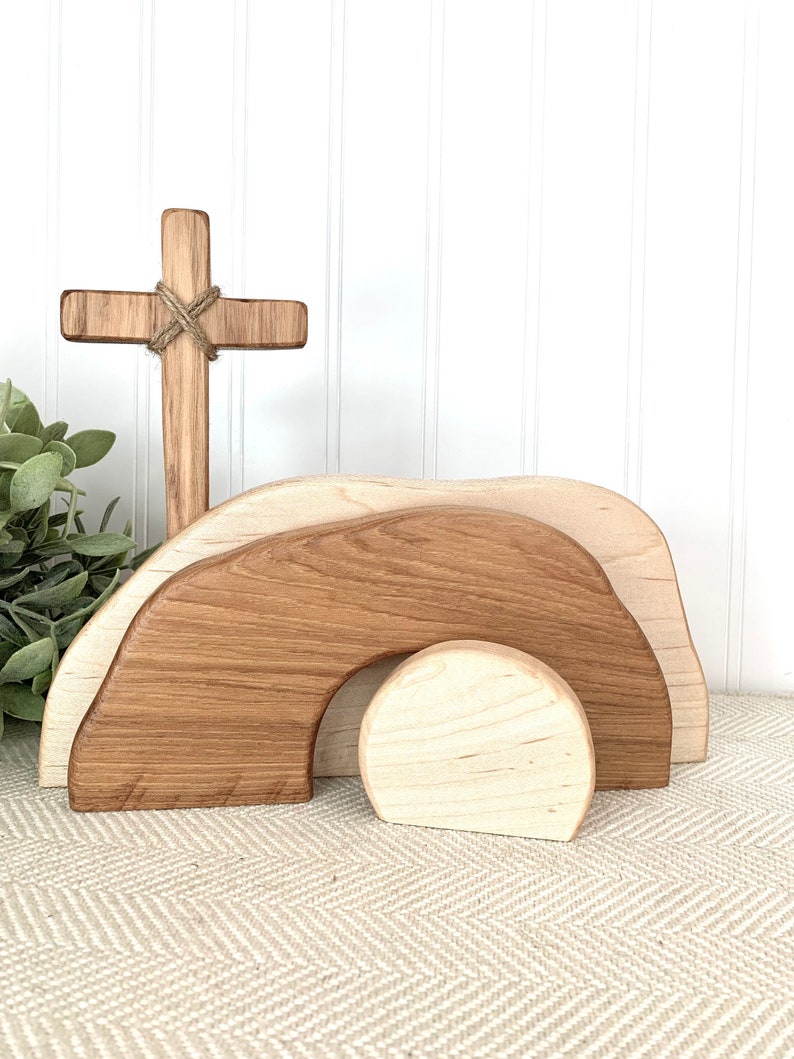 May include: Wooden Easter tomb set with a cross. The tomb is made of three pieces, a large brown piece, a smaller brown piece, and a round white piece. The cross is made of brown wood and has a piece of twine tied around it.