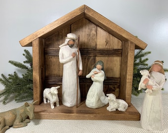 May include: A wooden nativity scene with white figurines of Mary, Joseph, and the baby Jesus. The scene also includes two white sheep and a shepherd with a staff. The scene is set against a brown wooden background.