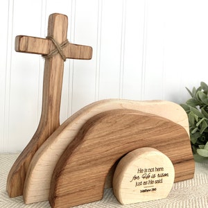 May include: Wooden Easter cross with a brown rope tied around it, standing in front of a wooden tomb with the inscription "He is not here. for He is risen, just as He said Mathew 286".