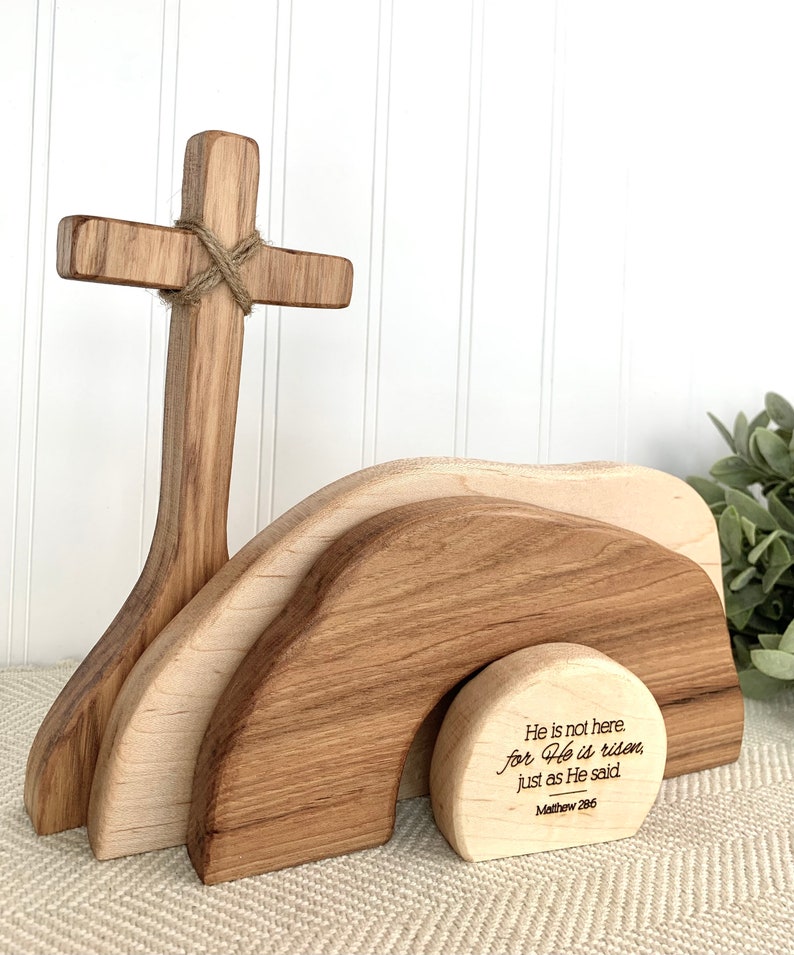 May include: Wooden Easter cross with a brown rope tied around it, standing in front of a wooden tomb with the inscription "He is not here. for He is risen, just as He said Mathew 286".