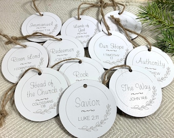 May include: A set of 11 round, white, wooden ornaments with black calligraphy and a sprig of leaves. Each ornament has a different Christian-themed phrase and a Bible verse. The phrases include "Immanuel", "Lamb of God", "Risen Lord", "Redeemer", "Head of the Church", "Rock", "Savior", "Our Hope", "Authority", "The Way", and "Ob"