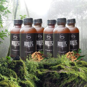 May include: Six brown bottles with black caps are arranged on a mossy surface. Each bottle has a black label with the word "ROOTS" in white, along with the "SACRED" logo and text. The background features a forest scene with green foliage.