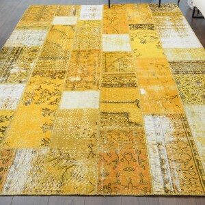Könnte beinhalten: Ein rechteckiger Patchwork-Teppich in Gelb-, Creme- und Brauntönen. Der Teppich weist verschiedene geometrische und florale Muster auf, die eine Vintage-Ästhetik erzeugen. Er liegt auf einem dunklen Holzboden.