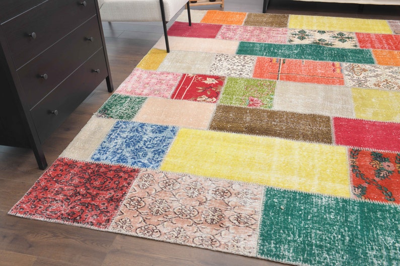 K&ouml;nnte beinhalten: Ein farbenfroher Patchwork-Teppich mit einer Vielzahl rechteckiger Formen in Rot-, Gelb-, Gr&uuml;n-, Blau- und Beiget&ouml;nen. Der Teppich hat ein abgenutztes, Vintage-Aussehen, das seinen einzigartigen Charakter unterstreicht. Der Teppich liegt auf einem Holzboden.