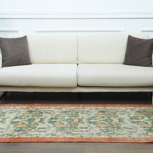 K&ouml;nnte beinhalten: Ein wei&szlig;es Sofa mit zwei braunen Kissen steht auf einem hellen Holzboden mit einem gemusterten Teppich davor. Der Teppich hat einen cremefarbenen Hintergrund mit orangefarbenen, gr&uuml;nen und blauen Blumendesigns. Ein wei&szlig;er Hocker steht rechts neben dem Sofa.