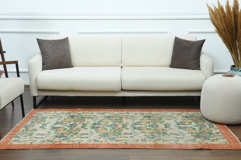 K&ouml;nnte beinhalten: Ein wei&szlig;es Sofa mit zwei braunen Kissen steht auf einem hellen Holzboden mit einem gemusterten Teppich davor. Der Teppich hat einen cremefarbenen Hintergrund mit orangefarbenen, gr&uuml;nen und blauen Blumendesigns. Ein wei&szlig;er Hocker steht rechts neben dem Sofa.