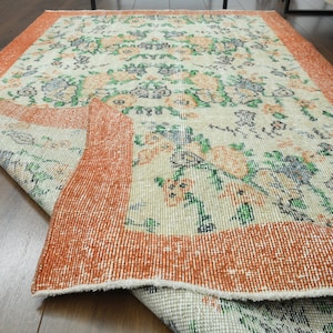 K&ouml;nnte beinhalten: Ein Vintage-Teppich mit einem floralen Muster in Orange-, Gr&uuml;n- und Wei&szlig;t&ouml;nen. Der Teppich hat einen ausgefransten Rand und einen abgenutzten Look.