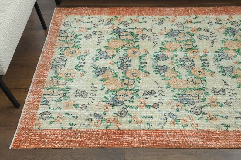 K&ouml;nnte beinhalten: Ein Vintage-Teppich mit einem floralen Muster in Orange-, Gr&uuml;n- und Blaut&ouml;nen auf cremefarbenem Hintergrund. Der Teppich hat einen rostfarbenen Rand.