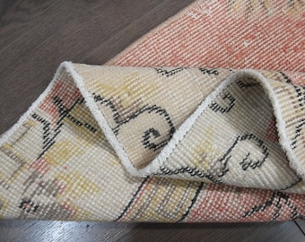 May include: A woven rug featuring a folded corner that reveals a cream-colored underside with black geometric patterns. The top surface displays a faded coral-red hue with intricate, repeating motifs in black, yellow, and cream throughout the fabric.