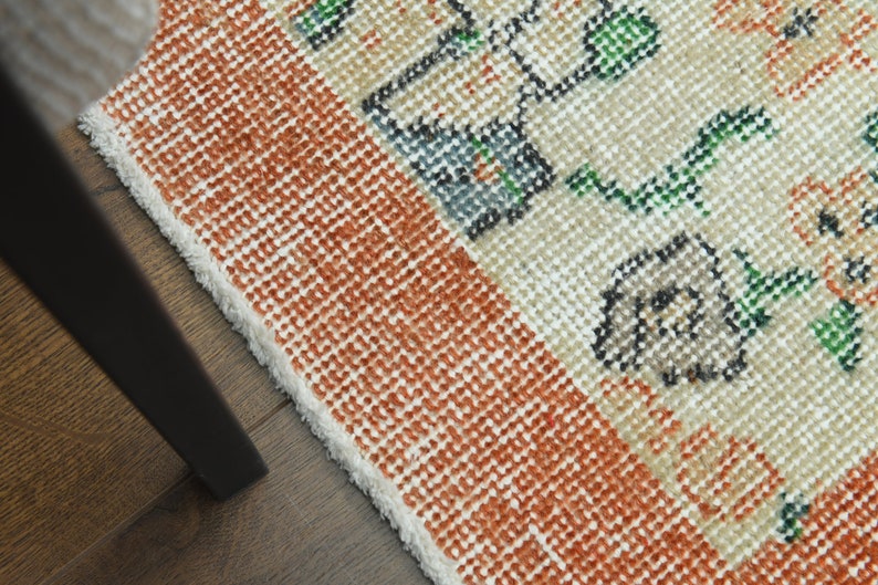 K&ouml;nnte beinhalten: Nahaufnahme eines gewebten Teppichs mit wei&szlig;em Hintergrund und orangefarbenen, gr&uuml;nen und schwarzen Blumenmustern. Der Teppich hat einen Fransenrand.