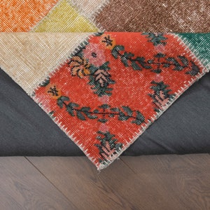 K&ouml;nnte beinhalten: Ein mehrfarbiger Patchwork-Teppich mit einer Vielzahl von Mustern. Der Teppich hat einen zentralen Bereich mit rotem Hintergrund und floralen Designs. Weitere Bereiche sind orange, beige, gelb, gr&uuml;n und braun. Der Teppich liegt auf einer dunkelgrauen Oberfl&auml;che.