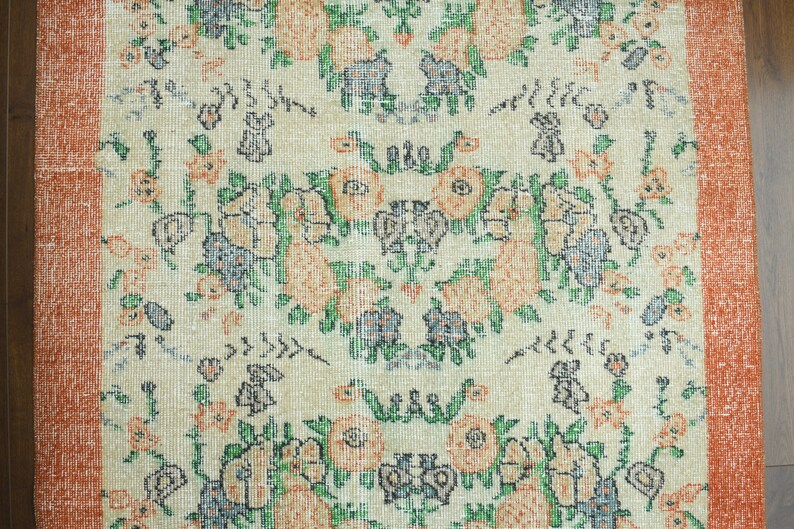 K&ouml;nnte beinhalten: Ein Vintage-Teppich mit cremefarbenem Hintergrund und einem floralen Muster in Orange-, Gr&uuml;n- und Blaut&ouml;nen. Der Teppich hat einen orangefarbenen Rand.