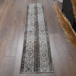 May include: A narrow, rectangular rug with a floral and striped design in grey and black tones. The rug is laid on a wooden floor. The design includes a central floral pattern with vertical stripes on either side. The rug is suitable for a hallway.