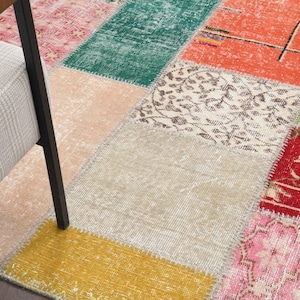 K&ouml;nnte beinhalten: Ein Patchwork-Teppich mit verschiedenen Farben und Mustern. Der Teppich besteht aus rechteckigen Teilen in Rosa, Gr&uuml;n, Orange, Beige und Gelb. Ein Stuhl mit hellem Sitz und dunklen Beinen ist teilweise links zu sehen.
