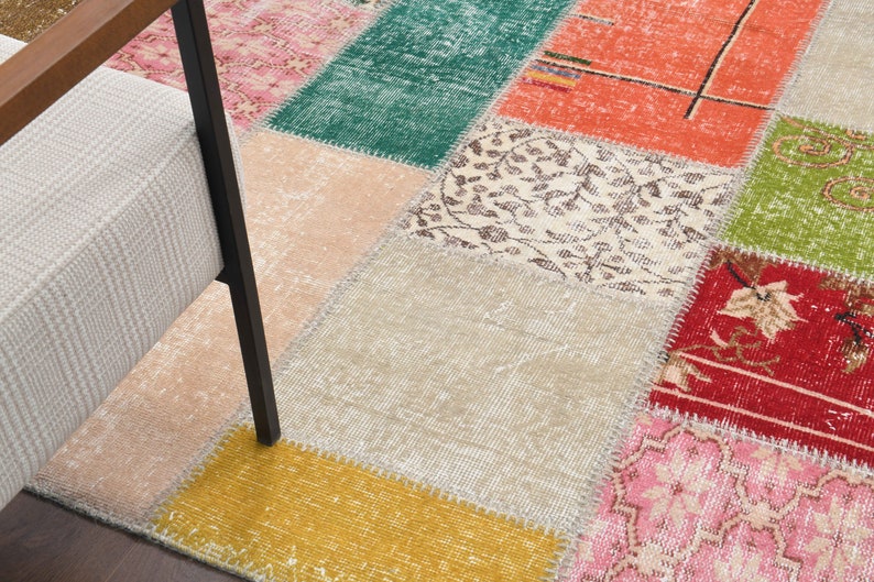 K&ouml;nnte beinhalten: Ein Patchwork-Teppich mit verschiedenen Farben und Mustern. Der Teppich besteht aus rechteckigen Teilen in Rosa, Gr&uuml;n, Orange, Beige und Gelb. Ein Stuhl mit hellem Sitz und dunklen Beinen ist teilweise links zu sehen.