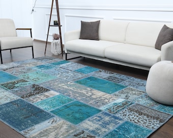 May include: A patchwork rug with a blue and gray color scheme. The rug is made up of different pieces of fabric sewn together, creating a unique and eclectic look. The rug is shown in a living room setting, with a white sofa and a gray ottoman.