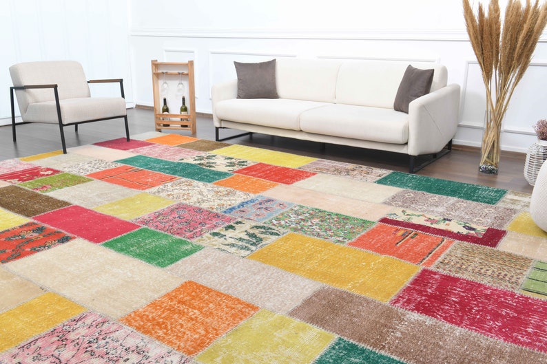 K&ouml;nnte beinhalten: Ein farbenfroher Patchwork-Teppich mit rechteckigen und quadratischen Mustern in Rot-, Gelb-, Gr&uuml;n- und Beiget&ouml;nen. Der Teppich befindet sich in einem Wohnzimmer mit einem wei&szlig;en Sofa und Stuhl.