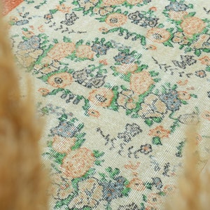 K&ouml;nnte beinhalten: Ein wei&szlig;er Teppich mit einem floralen Muster in Orange-, Gr&uuml;n- und Blaut&ouml;nen. Der Teppich hat einen Vintage-Look und -Feel.