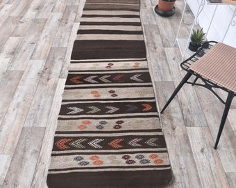 May include: A long, narrow rug with a brown and white geometric pattern. The rug has a fringe on both ends and is made of a woven material.