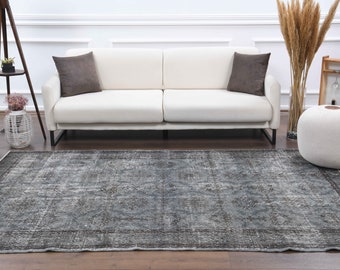 May include: A vintage-style rug with a faded blue and gray color scheme. The rug has a distressed look and features a geometric pattern. The rug is laid out on a wooden floor in front of a white sofa.