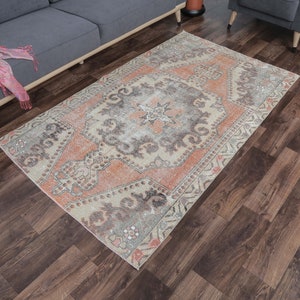 Puede incluir: Una alfombra vintage con un aspecto descolorido y desgastado. La alfombra tiene un medallón central con un diseño floral en tonos melocotón, gris y crema. La alfombra está rodeada por un borde con un patrón geométrico.