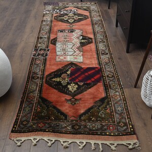 Puede incluir: Una alfombra de pasillo vintage con un diseño de patchwork. La alfombra tiene un esquema de color rojo, naranja y marrón con patrones intrincados. La alfombra está hecha de lana y tiene un borde de flecos.